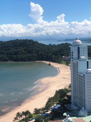 A coastal hotel with a beach view, a large building with a dome, and a forested hill in the background.