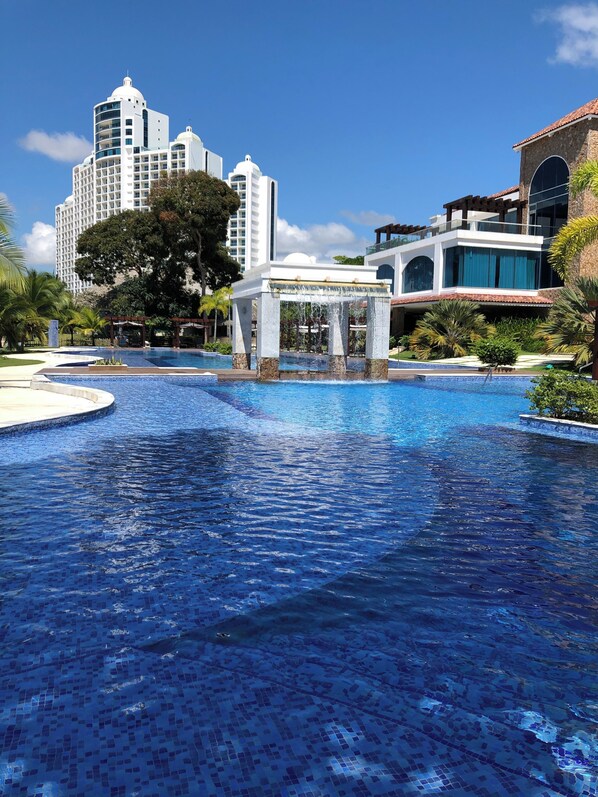 A swimming pool with a water fountain, surrounded by palm trees and modern buildings.