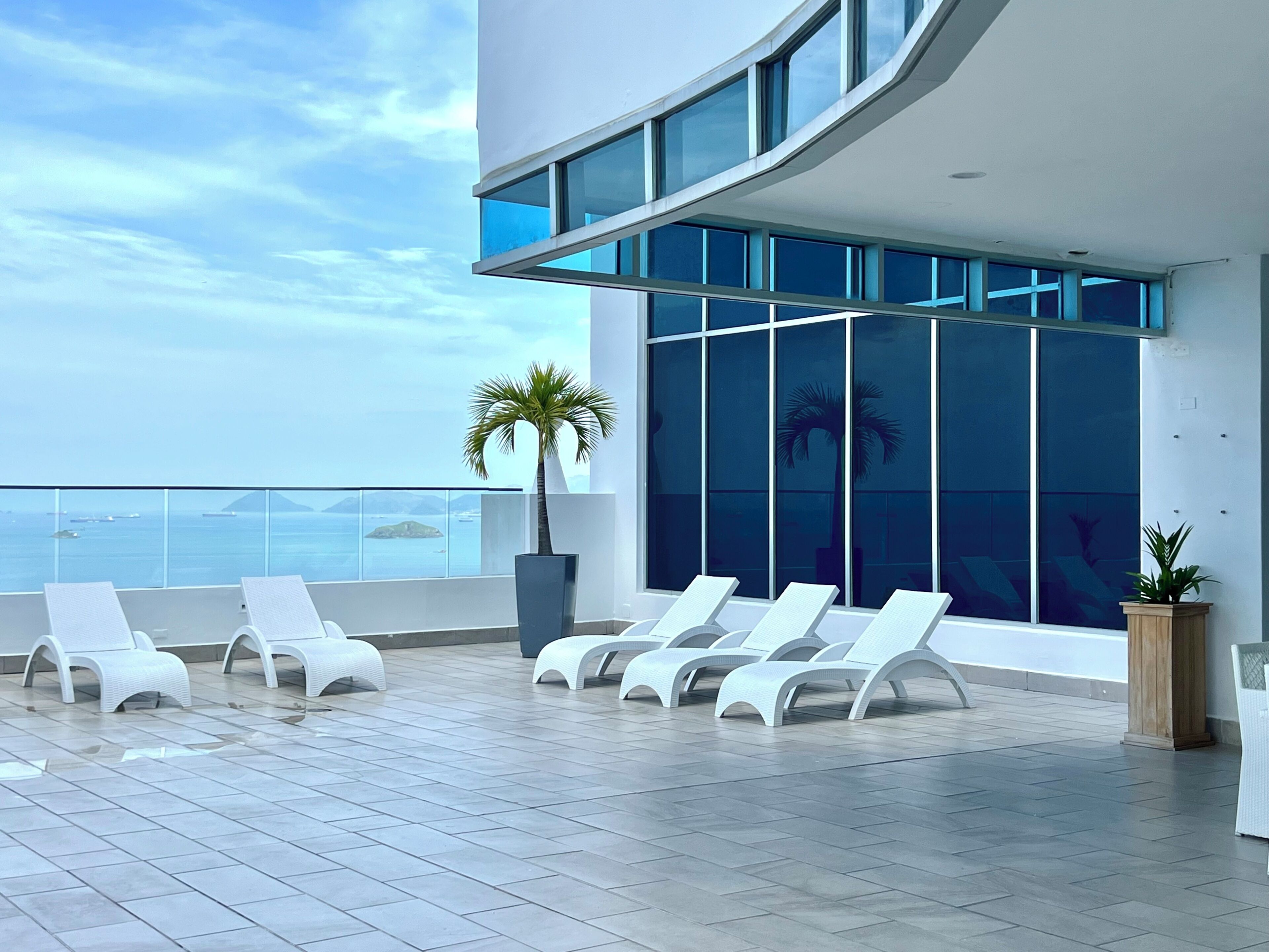 A modern rooftop terrace with white lounge chairs, large windows, and a view of the sea.