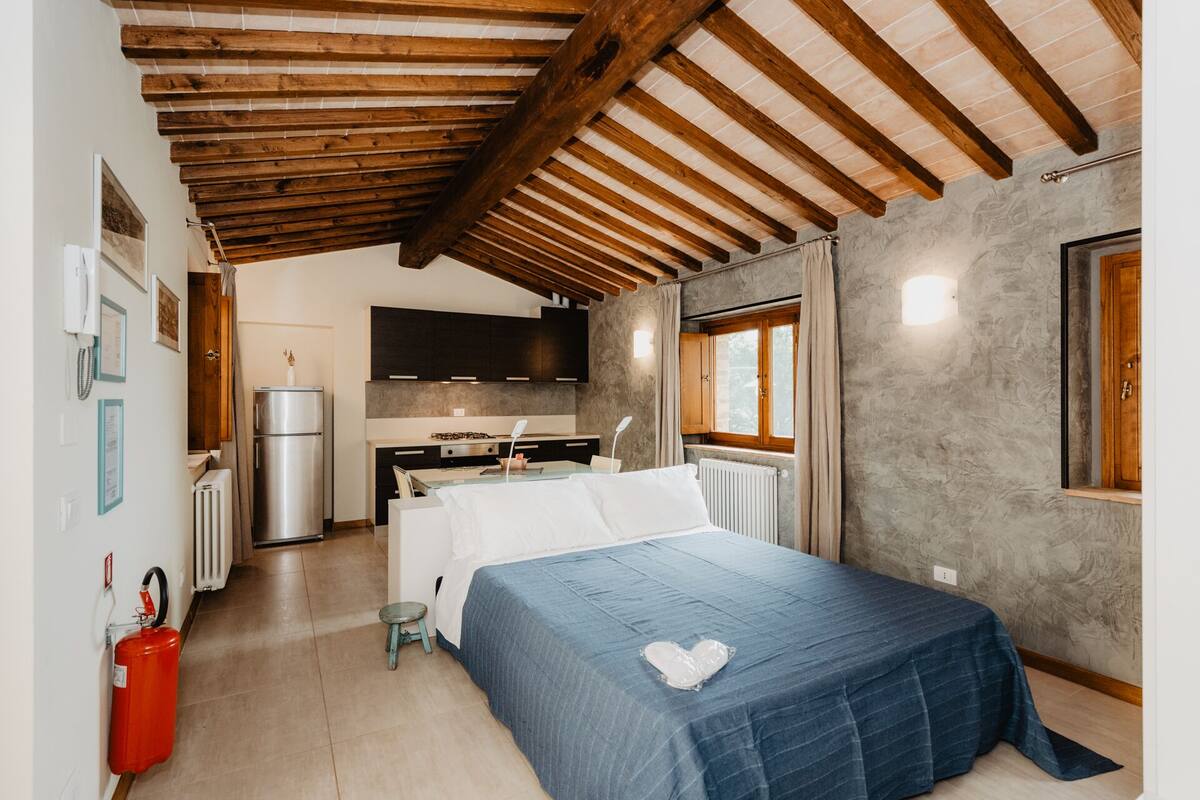 A bedroom with a bed, a small stool, a red fire extinguisher, a kitchen area with a stove, and a window with curtains.