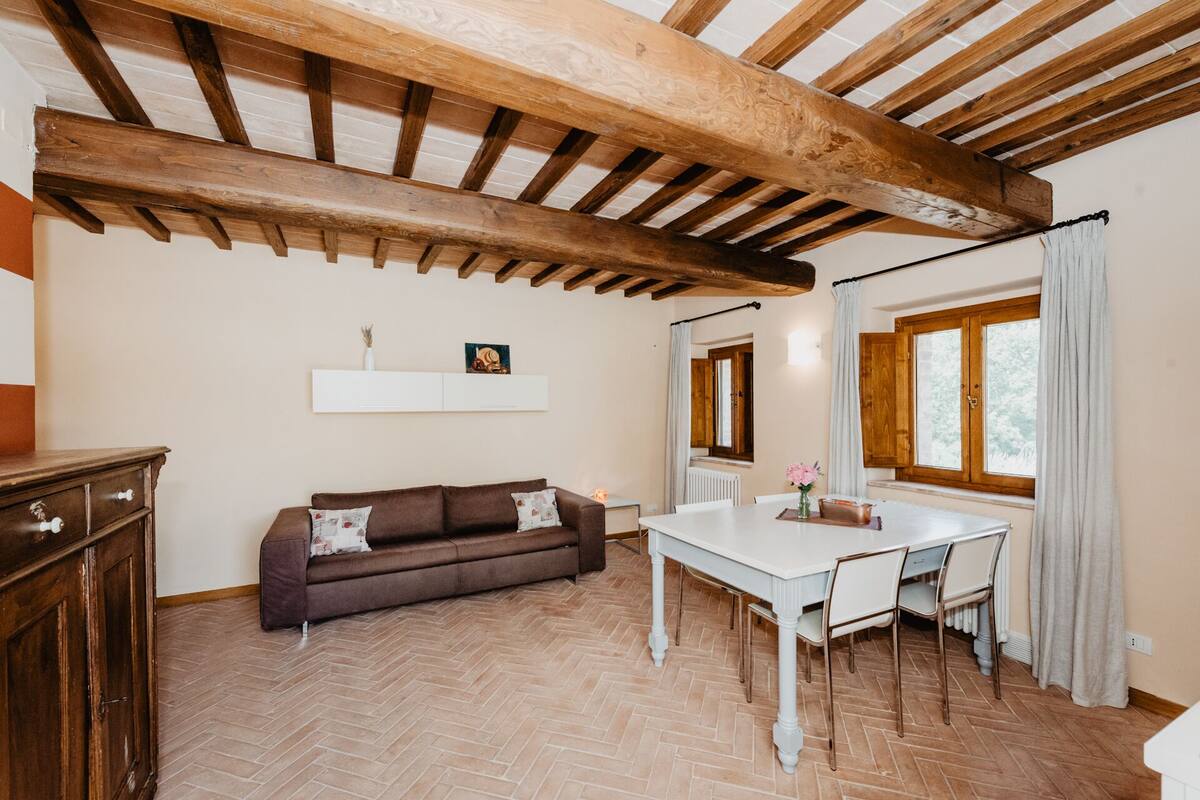 A room with a herringbone floor, wooden ceiling beams, a brown sofa, a white table with chairs, a window with curtains, and a radiator.