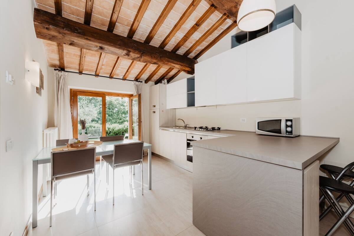 A modern kitchen with a central island, a dining area with chairs, and a view of greenery outside.