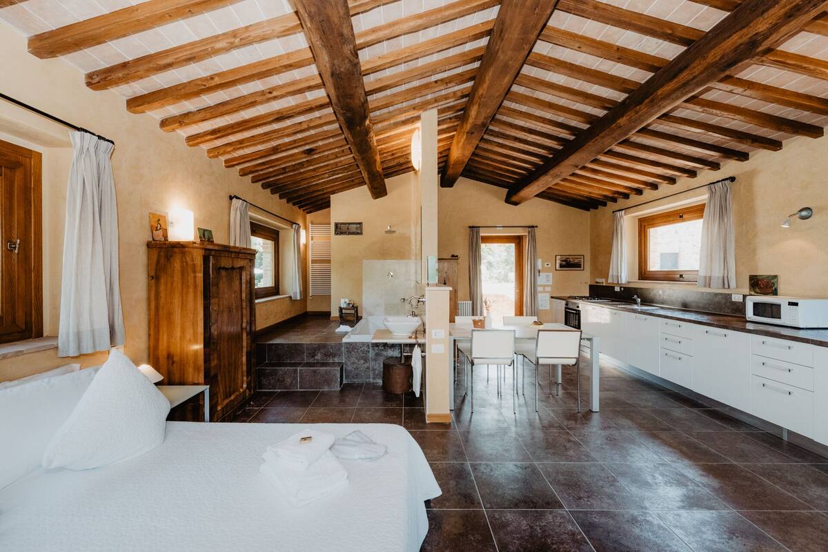 A spacious room with a wooden ceiling, a bed with white linens, a bathroom with a sink and mirror, and a kitchen with white cabinets and a microwave.