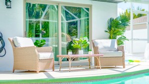 Ein Poolbereich mit Korbstühlen und einem Tisch, eine Glastür, die zu einem Garten führt, und freier Blick auf den Pool.