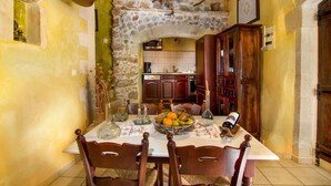 Un coin repas agrémenté d’une arche en pierre, de chaises en bois, d’une table sur laquelle se trouve un panier de fruits et une bouteille de vin.