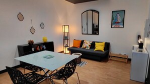 A modern living room with a glass dining table, black chairs, a black sofa with colorful cushions, a tall floor lamp, a refrigerator, and a mirror on the wall. - Apartment in the Rotonde district (Vichy)