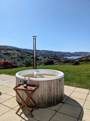 A hot tub with a wooden exterior and a metal chimney, set on a patio with a scenic view of hills and a body of water. - Beautifully positioned farmhouse overlooking hills and sea (Oban, Lerags)
