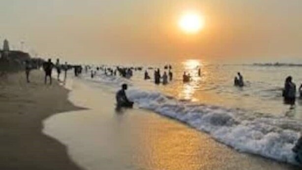 Escena de playa con el sol poniéndose sobre el agua, gente en el agua y una orilla arenosa.