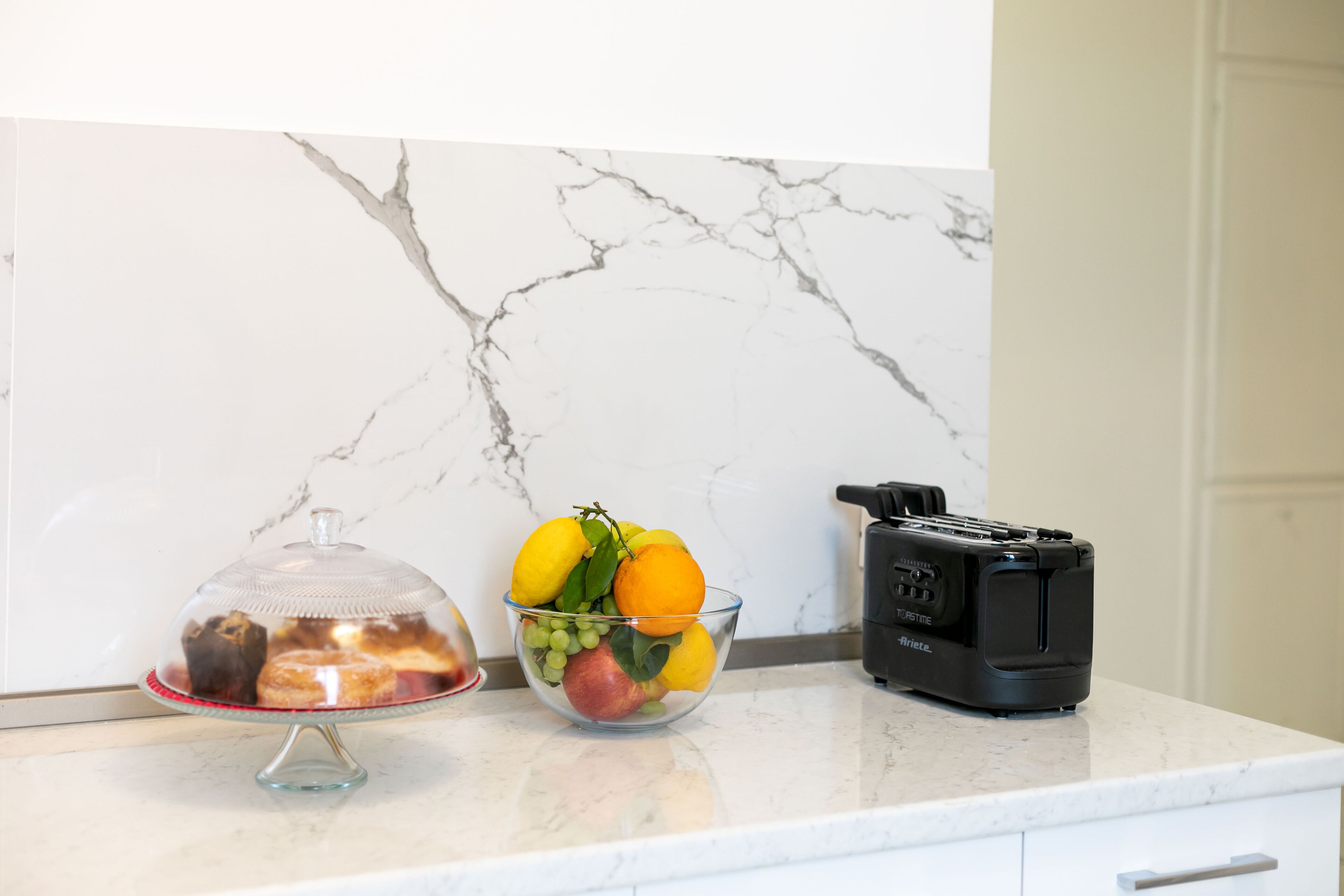 A kitchen counter with a marble backsplash, a glass bowl of fruit, a covered dessert, and a toaster.