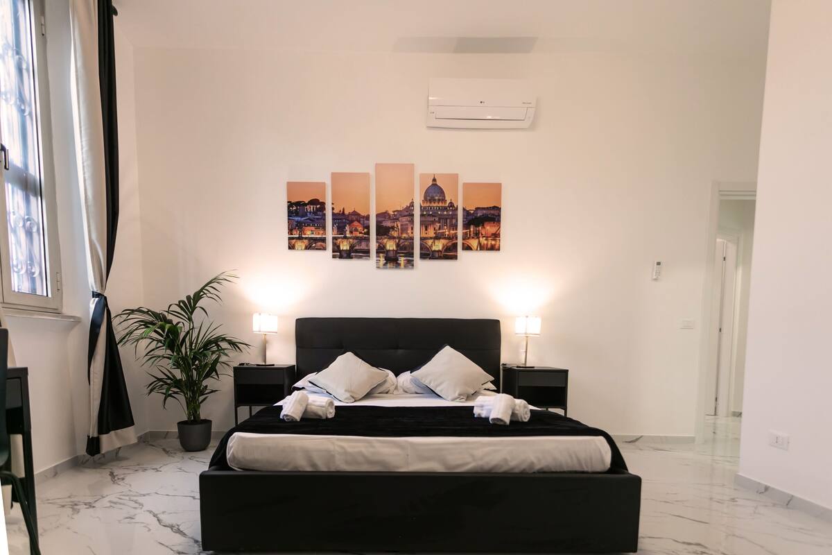 Moderni hotellihuone, jossa on musta sänky, valkoiset verhot, ruukkukasvi ja kaupunkimaisemaa esittävä taideteos seinällä.