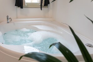 Ett badkar fyllt med vatten och bubblor, ett fönster ovanför och en växt i förgrunden.