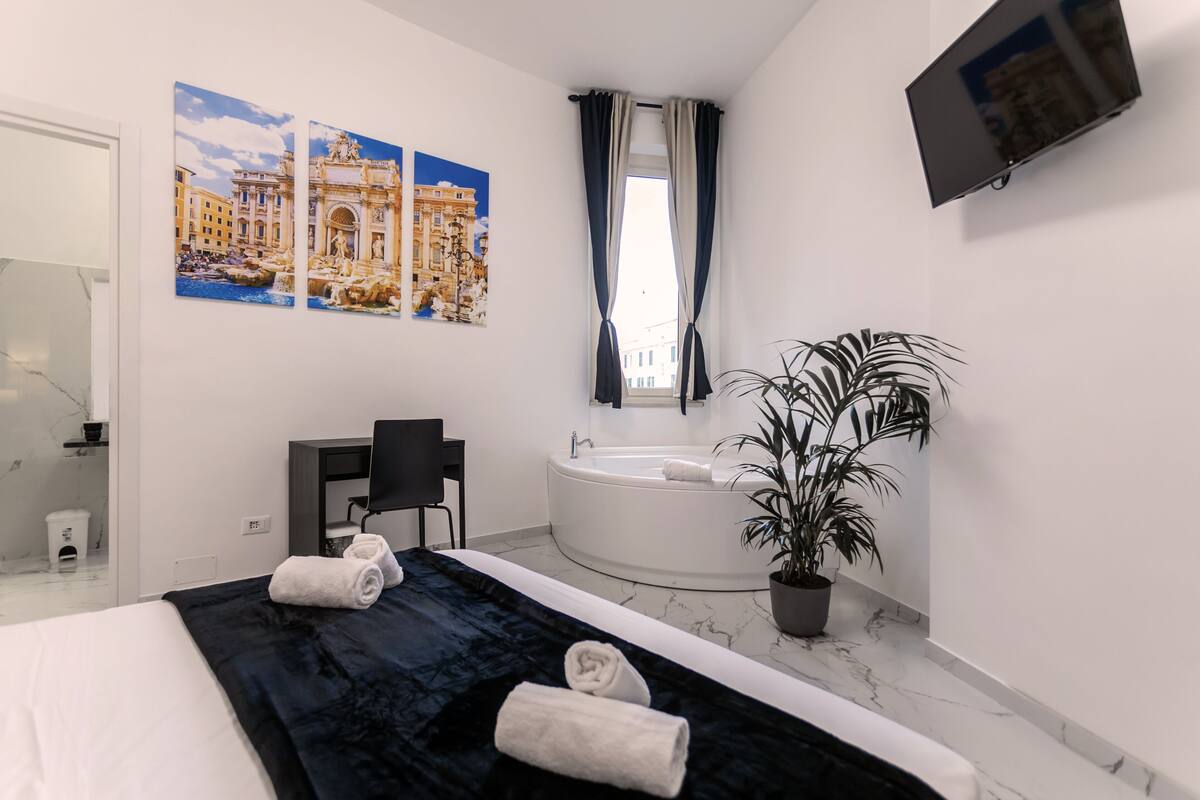 Moderni hotellihuone, jossa on suuri sänky, musta heittopeitto, kaksi rullalle taitettua pyyhettä, musta tuoli, ruukussa oleva kasvi, suuri ikkuna verhoilla, taulutelevisio ja seinälle ripustettu maalaus, jossa on kuvattuna historiallinen rakennus.