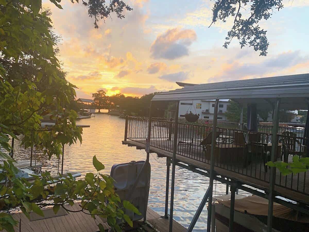 A riverside deck with a view of the sunset, a dock with boats, and a building across the water.