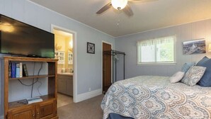 Un dormitorio con una cama, un televisor sobre un soporte de madera, un ventilador de techo y una ventana con cortinas.