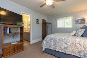A bedroom with a bed, a television on a wooden stand, a ceiling fan, and a window with curtains.