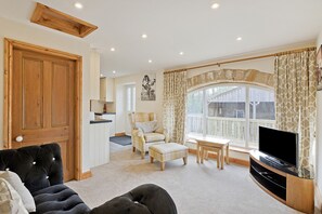 A living room with a black sofa, a wooden TV stand, a dining table, and a large window with curtains. - Tom's Cabin (Knaresborough)