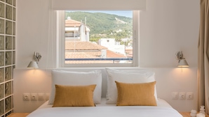 A hotel room with a bed, two bedside lamps, a window with a view of buildings and hills, and a wall with a glass block pattern. - Aria Blue Suites (Skiathos)