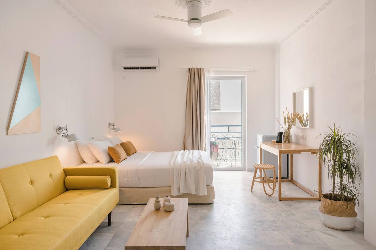 Moderni hotellihuone, jossa on keltainen sohva, valkoisilla lakanoiden päällä varustettu sänky, puinen työpöytä ja ruukussa oleva kasvi.