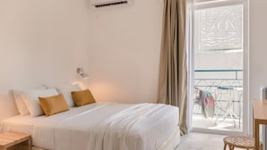 A bedroom with a bed, bedside table, and a view of a balcony with a railing and a chair. - Aria Blue Suites (Skiathos)