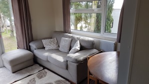 A cozy living room with a grey sofa, two white cushions, a wooden chair, and a window with a view of trees.
