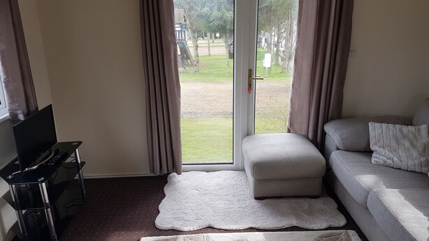 A room with a sofa, a small ottoman, a TV on a stand, and a view of a grassy area through a glass door.