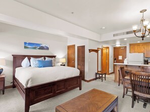 A hotel room with a large bed, a wooden headboard, a bedside table with a lamp, a small desk, a chair, a wooden coffee table, and a view of the kitchen area. - Sundial B404 | Ski-in/Ski-out | Mountain Views | Full Kitchen | Pools & Hot Tubs (Park City)
