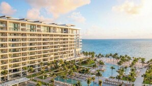 A hotel with multiple floors, a pool area with palm trees, and a view of the ocean.