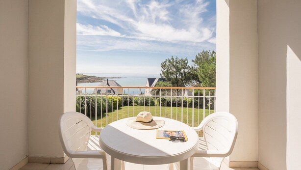 A balcony with a white table and two white chairs, overlooking a coastal view with houses and greenery. - Residence Pierre & Vacances Audierne le Phare de Trescadec (Audierne)