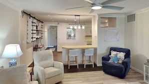 Un salon comprenant un canapé, un fauteuil et une chaise bleue. On y trouve également un coin repas avec une table et des chaises. Un ventilateur de plafond est visible.