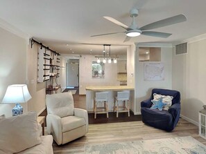A living room with a sofa, armchair, and a blue chair. There is a dining area with a table and chairs. A ceiling fan is visible. - Hibiscus 102-D by Distinctive Beach Rentals (St. Augustine)
