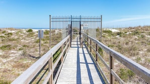 Une promenade en bois mène à une entrée fermée par un portail, d’où l’on a une vue dégagée sur la plage et l’océan.
