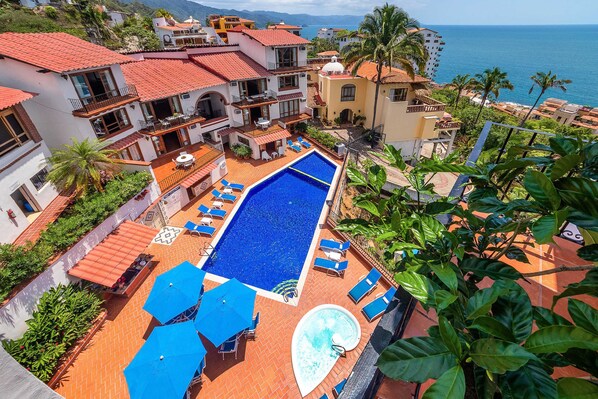 A rooftop pool area with multiple lounge chairs and umbrellas, surrounded by buildings with red-tiled roofs and balconies overlooking the sea.