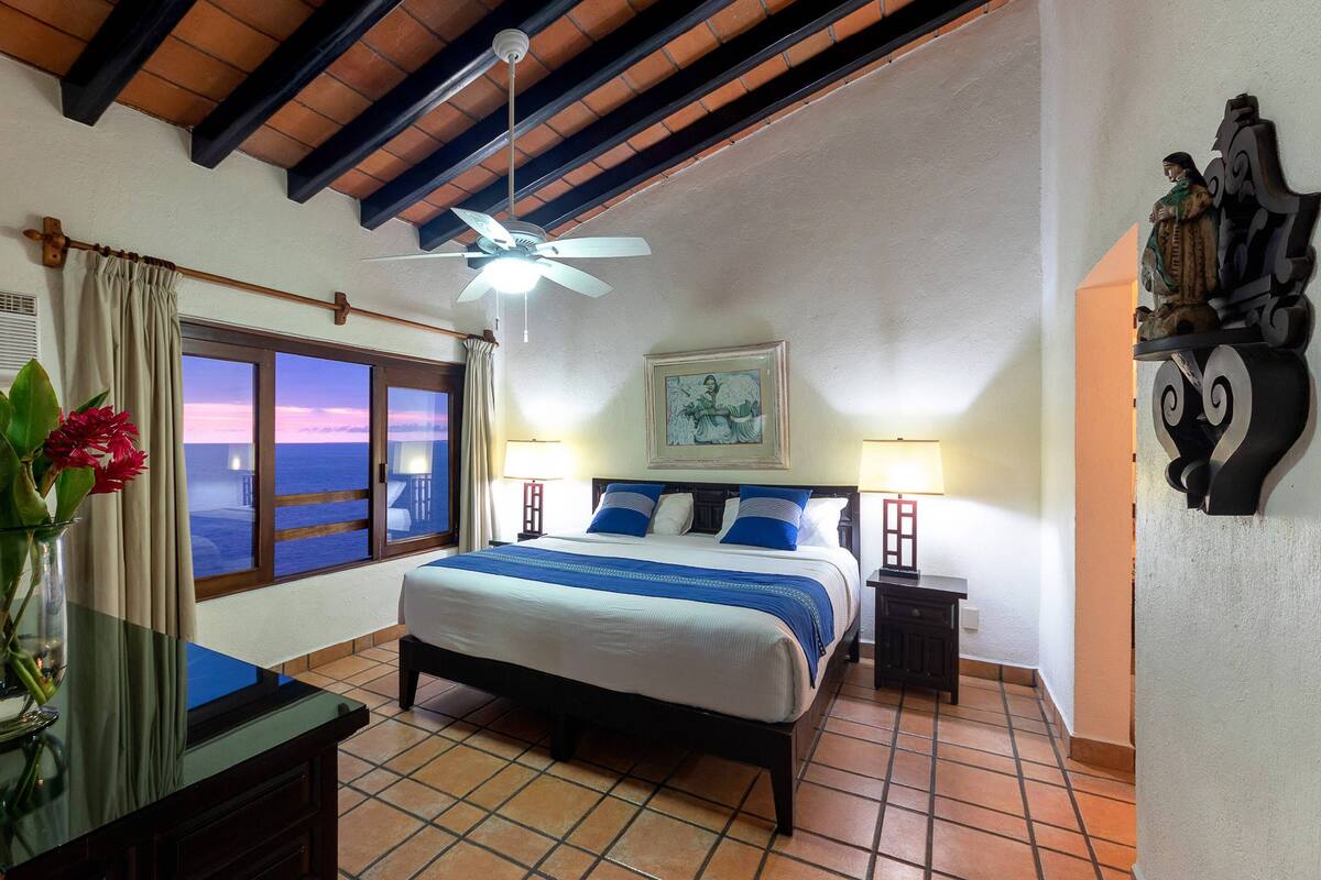 A hotel room with a bed, a nightstand, a desk, a window with a view of the ocean, a ceiling fan, and a painting on the wall.