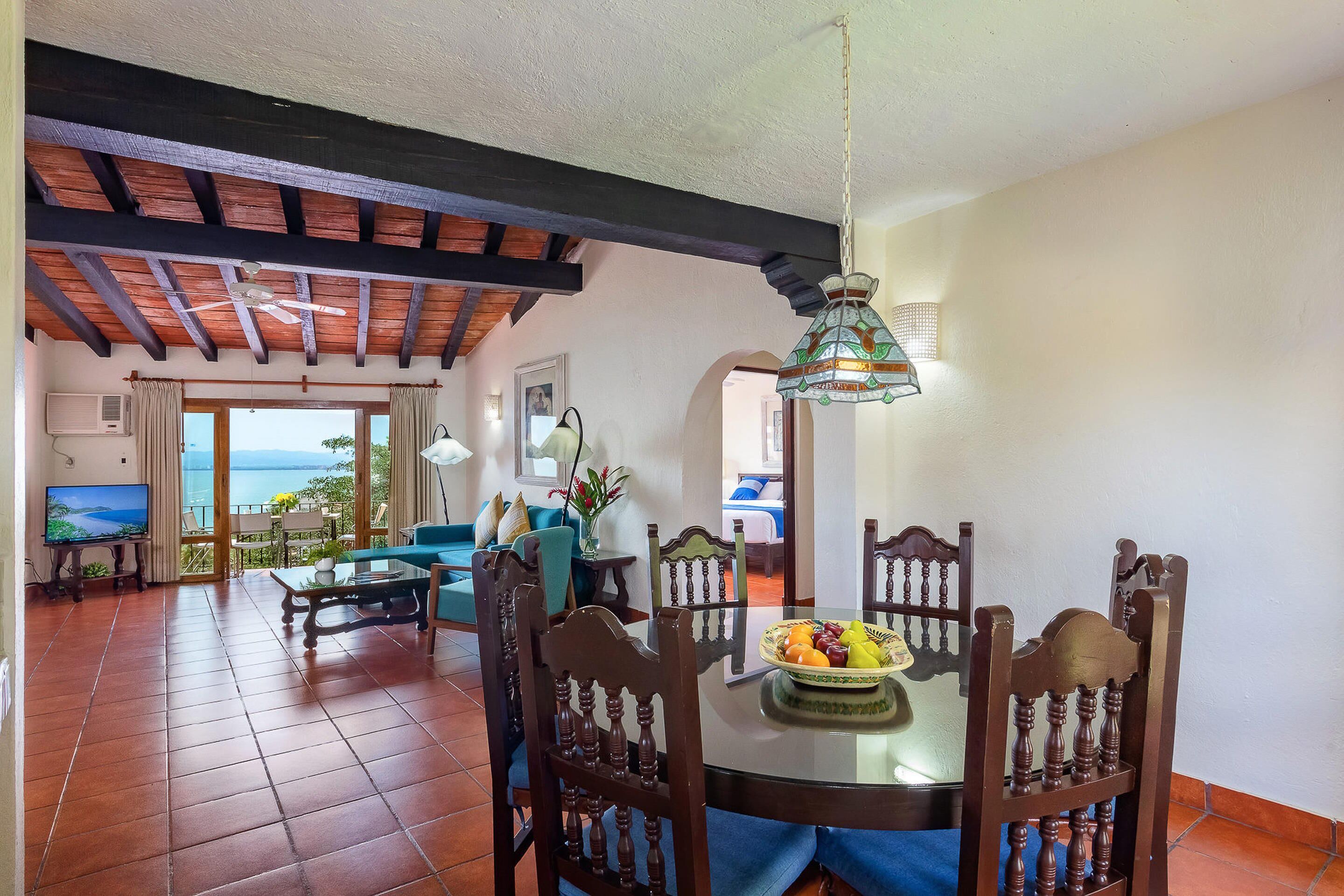 A dining area with a round table, chairs, and a fruit bowl. There is a television and a view of the sea through large windows.