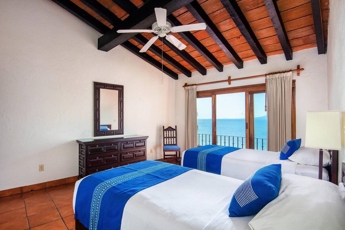 A hotel room with two beds, a ceiling fan, a dresser, a chair, and a view of the sea.