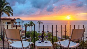 Ein Tisch mit zwei Stühlen auf einem Balkon mit Blick auf das Meer, inklusive Sonnenuntergang.