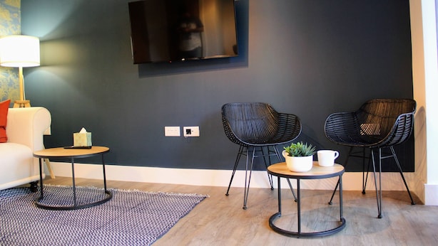 Una sala moderna con un televisor de pantalla plana montado en la pared, dos sillas negras, una mesita redonda con una planta y una taza, y un sofá blanco con un cojín rojo.