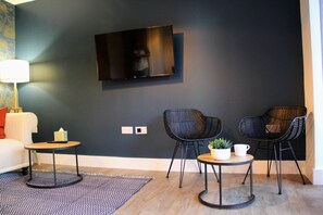 A modern living room with a flat-screen TV mounted on the wall, two black chairs, a small round table with a plant and a cup, and a white sofa with a red pillow. - 3 Double Bed Luxurious House Near Heathrow Airport (Staines)