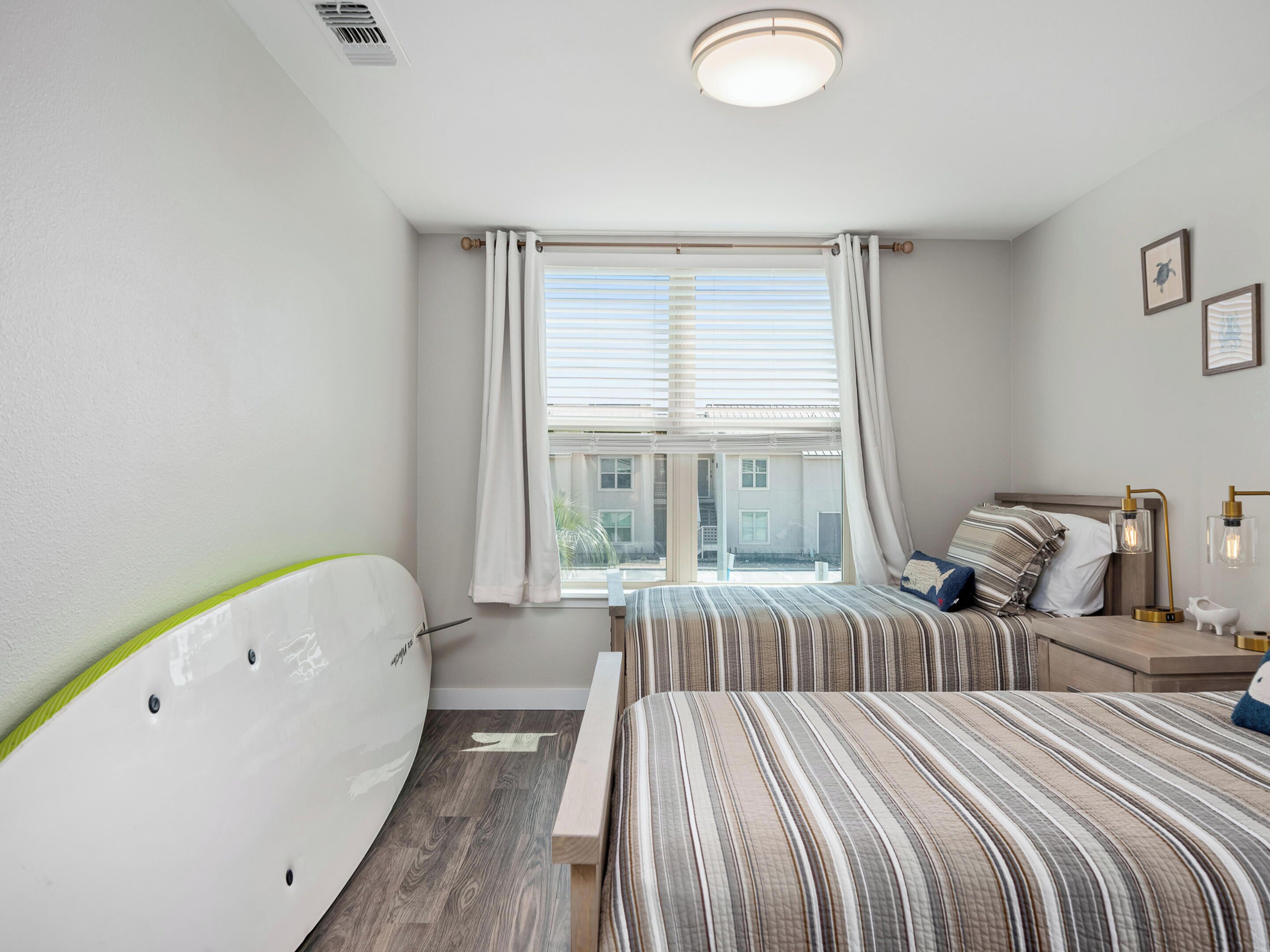 Ein Schlafzimmer mit zwei Betten, einem Fenster mit Vorhängen, einem an der Wand angelehnten Surfbrett und einer Deckenleuchte.
