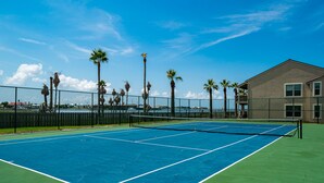 An outdoor tennis court with a net, surrounded by a fence and palm trees, with a building in the background. - Little's Toasty Coasty by Miss Kitty’s (Rockport)