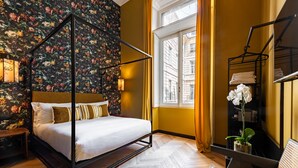 Un lit à baldaquin avec un papier peint à motifs floraux, un parquet en bois disposé en chevrons, une fenêtre avec des rideaux jaunes et une table de chevet avec une lampe.