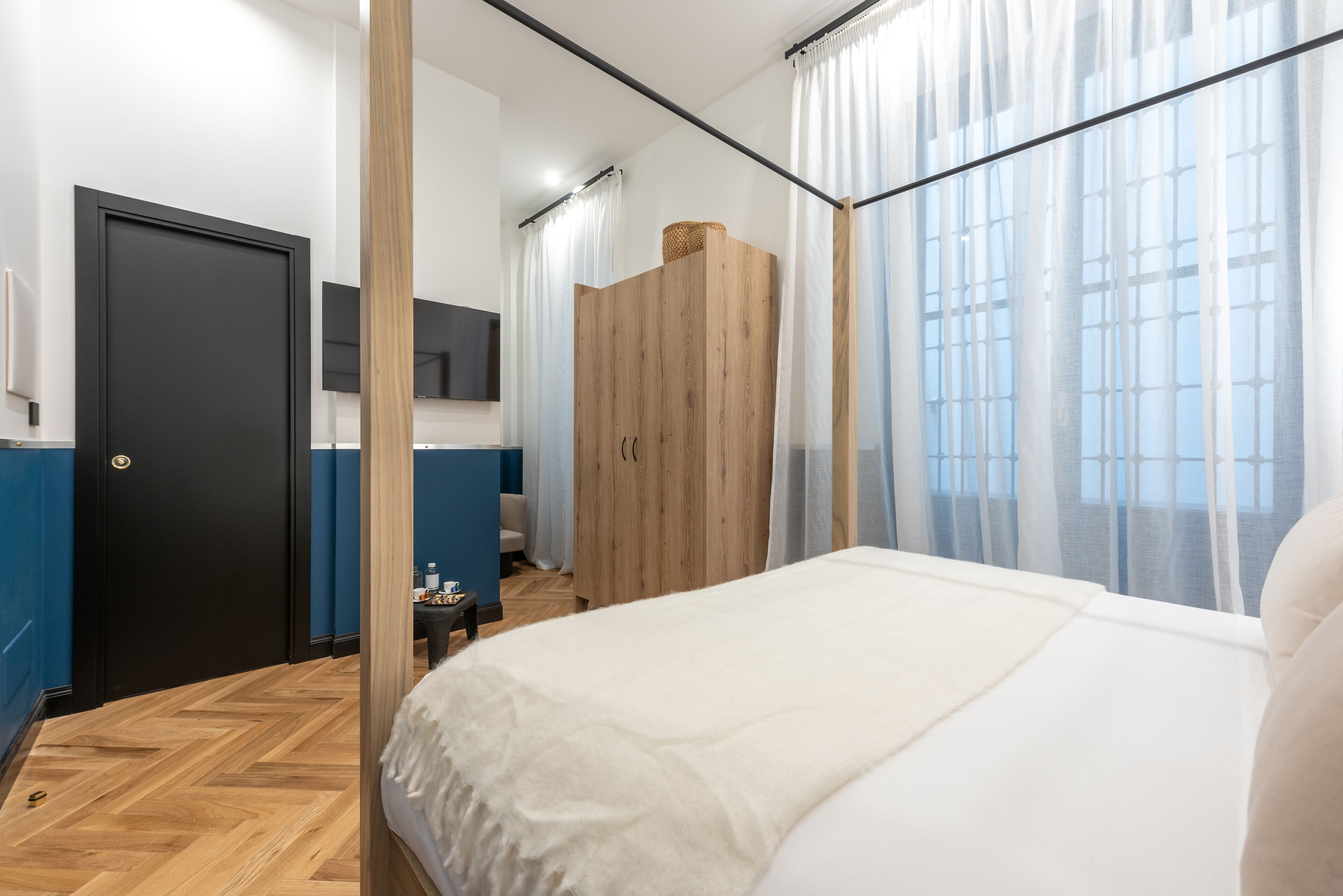 Ein modernes Schlafzimmer mit einem großen Bett, einem Holzschrank, einem kleinen Tisch mit einer Lampe und Blick auf die Stadt durch transparente Vorhänge.
