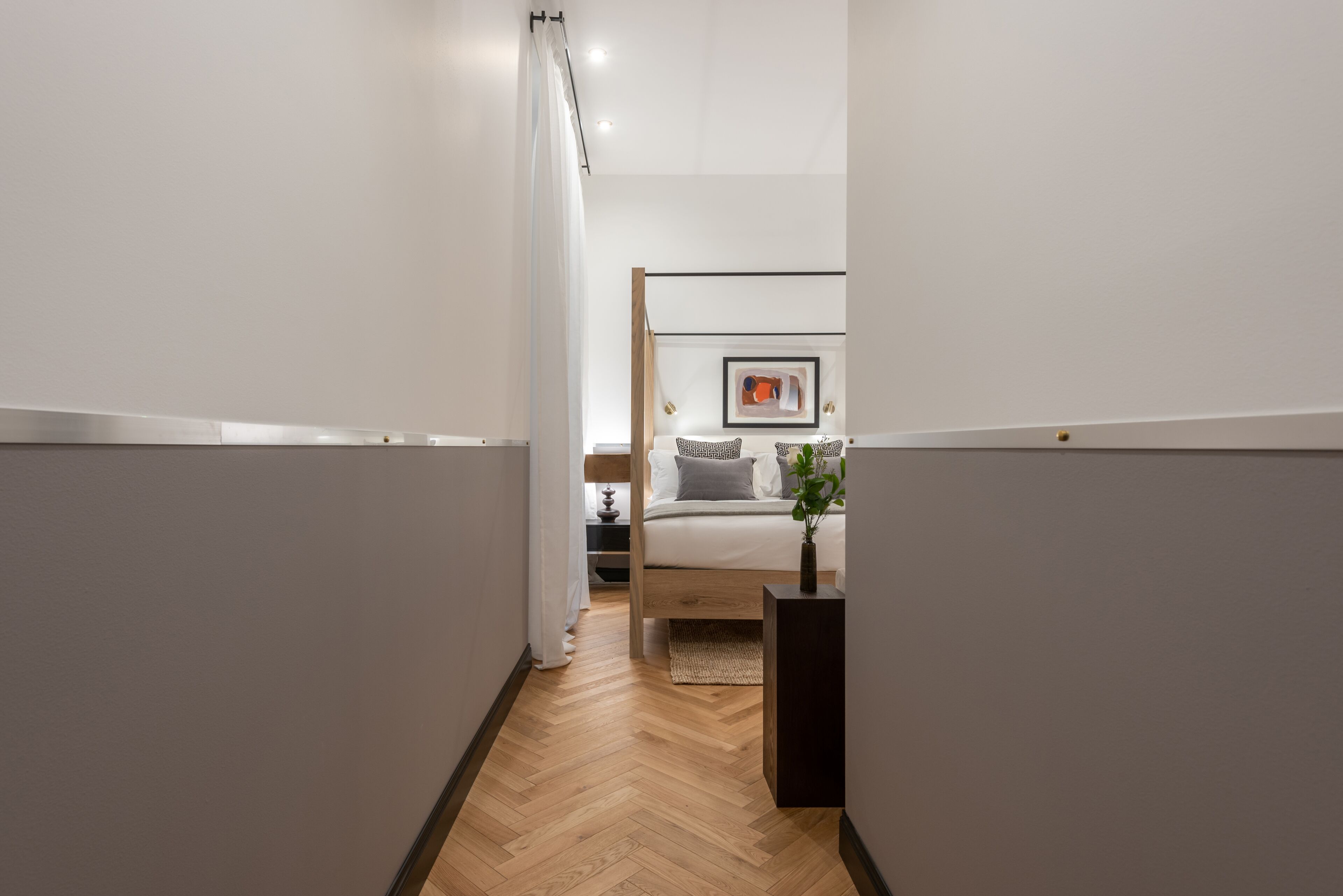 Un pasillo estrecho con suelo de madera que conduce a un dormitorio con cama, mesita de noche y un cuadro en la pared.