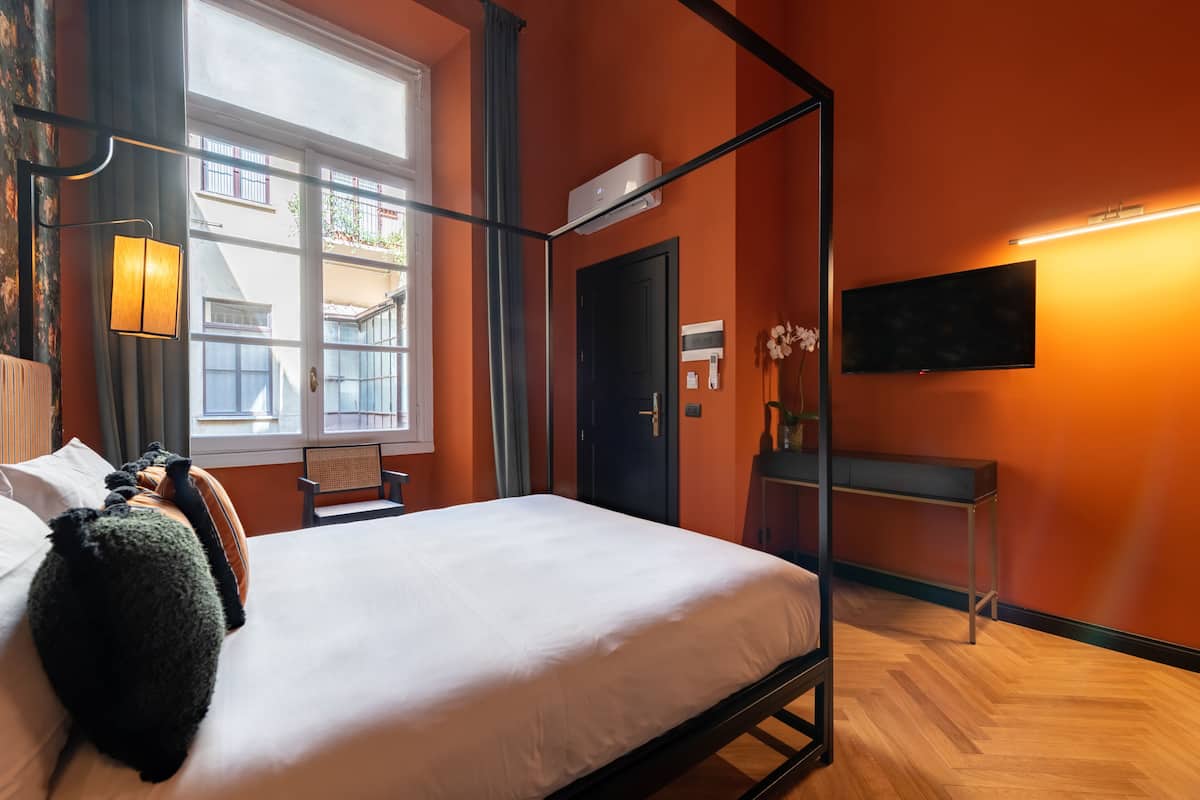 A hotel room with a four-poster bed, a flat-screen TV, a desk, and a window with a view of buildings.
