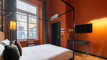 A hotel room with a four-poster bed, a flat-screen TV, a desk, and a window with a view of buildings.