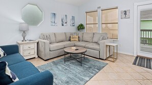 A living room with a sofa, coffee table, and a blue armchair. There is a ceiling fan, a lamp, and a window with blinds. - Sandy Pants Beach House by Miss Kitty’s (Rockport)