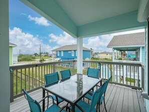 A covered outdoor area with a glass-top table and blue chairs, overlooking a residential neighborhood. - Sandy Pants Beach House by Miss Kitty’s (Rockport)