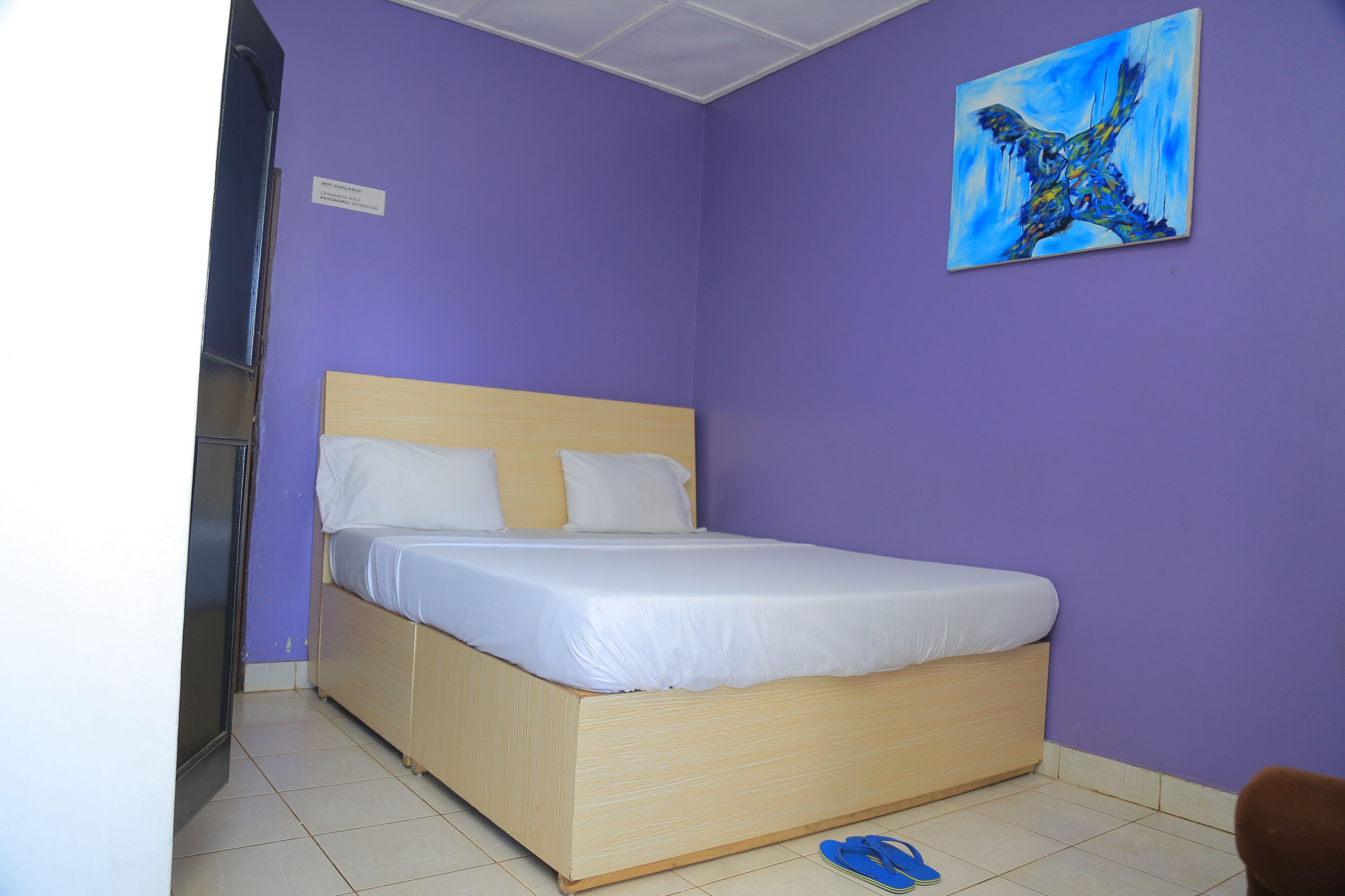 A bedroom with a bed, a painting on the wall, and a pair of slippers on the floor.
