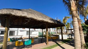 An outdoor seating area with a thatched roof, surrounded by palm trees and a white building in the background. - Alondra 3304 ByTrvl2hm (Mazatlán)