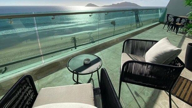 A balcony with a glass railing overlooking a beach and ocean. There are wicker chairs and a small round table with a cushion. - CONDOS ONE ByTrvl2hm & Mazatlan Regency (Mazatlán)